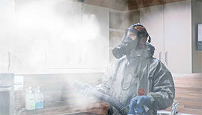 Ein Techniker mit Atemmaske, Schutzanzug und Handschuhen desinfiziert einen Raum.